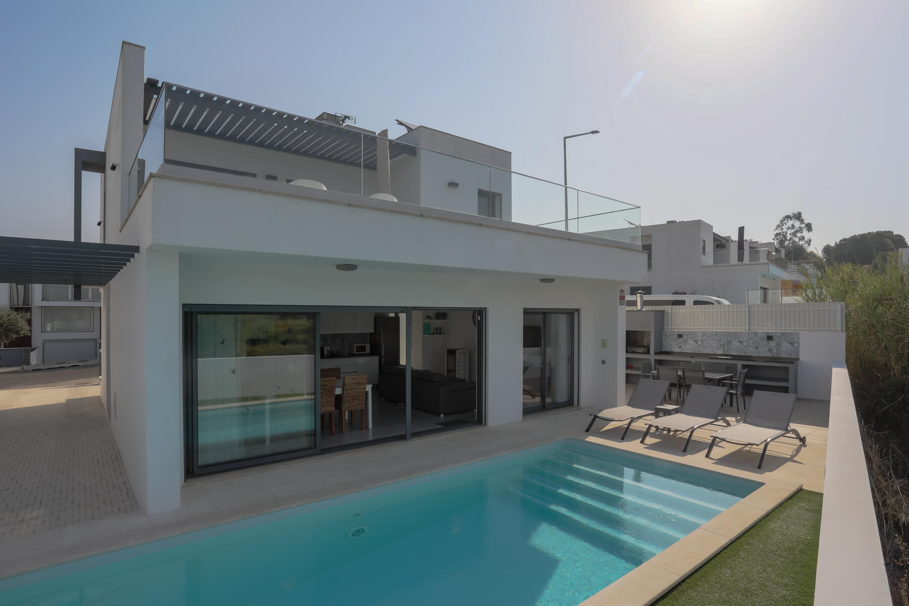Piscina exterior aquecida privativa com várias espreguiçadeiras, uma mesa com cadeiras e uma zona de barbecue, delimitada por uma vedação branca com vista para o jardim, na zona rústica de Coto, Caldas da Rainha.